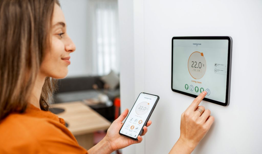 Woman using her smart heating system via a smart device built onto the wall and holding her phone with the smart heating app